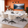 Modern boardroom featuring a high-gloss white writable conference table with a slightly reflective surface, showing handwritten notes and markers. The table is surrounded by five cream boucle swivel chairs on casters. A laptop sits open on the table. The room includes a burnt orange accent wall with a wall-mounted TV, a light wood sideboard, and multiple decorative plants, creating a warm, contemporary workspace.